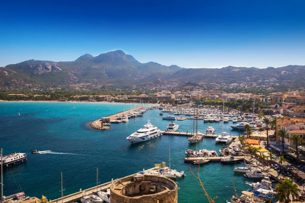 port-de-plaisance-de-calvi-vue-panoramique-bateaux-mer-corsica Vue aérienne du port de plaisance de Calvi, montrant des yachts et bateaux sous un ciel bleu, avec des montagnes en arrière-plan et des palmiers bordant la promenade.