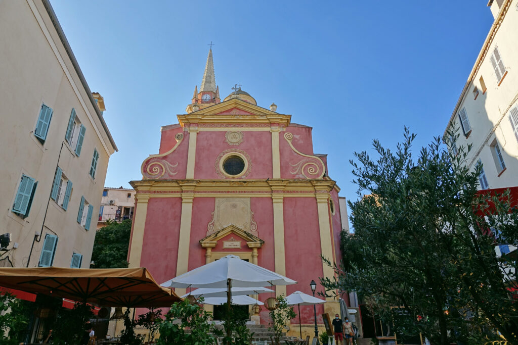 eglise-sainte-marie-majeure-dans-une-cour-coloree Église pittoresque Sainte-Marie-Majeure illuminée de rose et entourée de verdure, offrant une ambiance méditerranéenne.