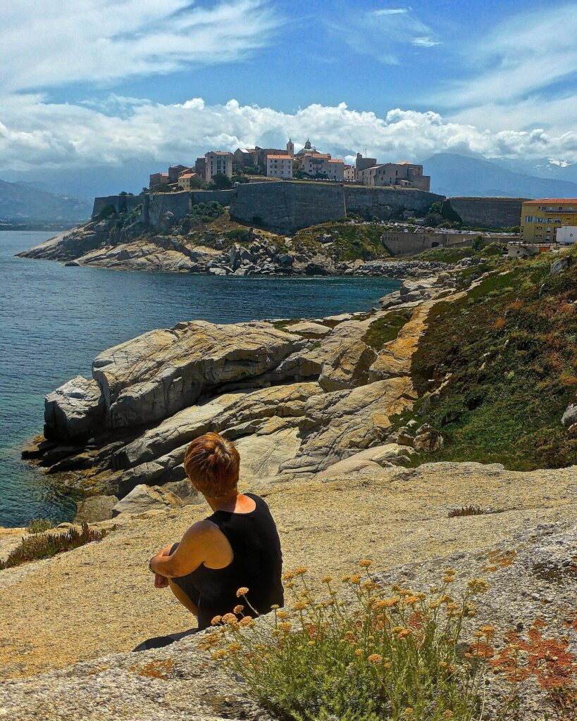 vue-panoramique-de-calvi-en-haute-corse-au-bord-de-la-mer Un homme contemplant l'horizon méditerranéen depuis les rochers de Calvi, entouré par une atmosphère de sérénité et de contemplation, sous le ciel changeant de Haute-Corse. Comment choisir un Camping Corse bord de mer à Calvi ?