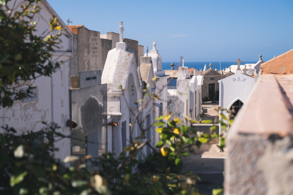 cimetiere-au-bord-de-la-mer-sous-un-ciel-clair Image d'un cimetière paisible au bord de la mer sous un ciel bleu, entouré de tombes blanches et de verdure, évoquant calme et tranquillité.