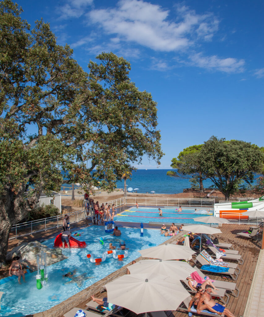 Une famille s'amuse dans une piscine en bord de mer entourée de palmiers et glisse sur des toboggans sous le soleil éclatant.