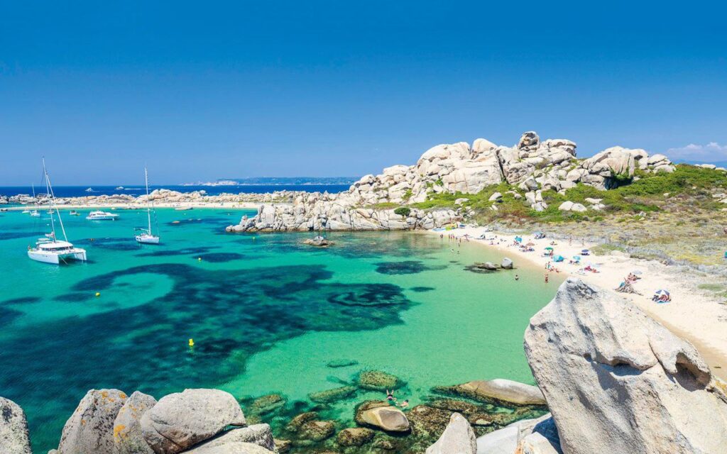 plage-de-corse-avec-sable-fin-et-eaux-cristallines Image d'une plage corse idyllique : sable doré, eaux turquoise et voiliers sous un ciel bleu éclatant.