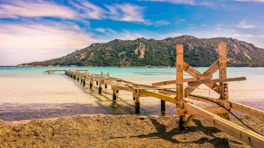pont-pittoresque-a-santa-giulia-en-corse Pont en bois sur la plage de Santa Giulia en Corse, sur fond de mer turquoise et collines verdoyantes, évoquant des vacances parfaites.