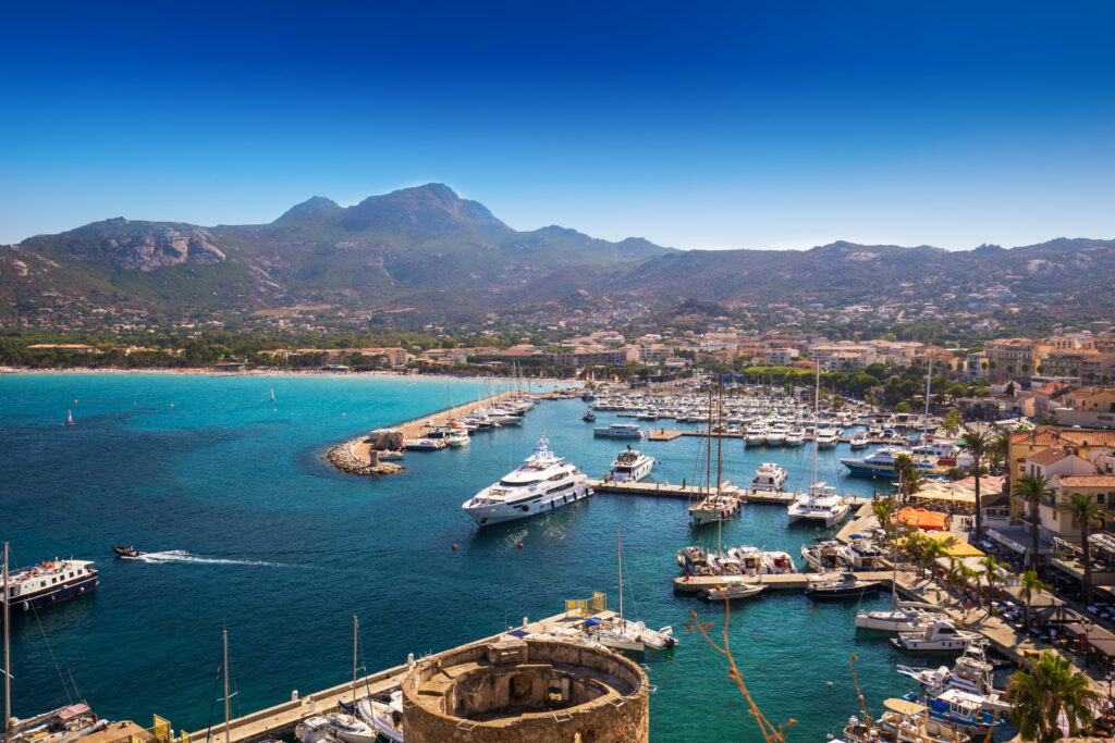 port-de-plaisance-de-calvi-sous-un-ciel-azur Vue aérienne du port de plaisance de Calvi, montrant des yachts et bateaux sous un ciel bleu, avec des montagnes en arrière-plan et des palmiers bordant la promenade.