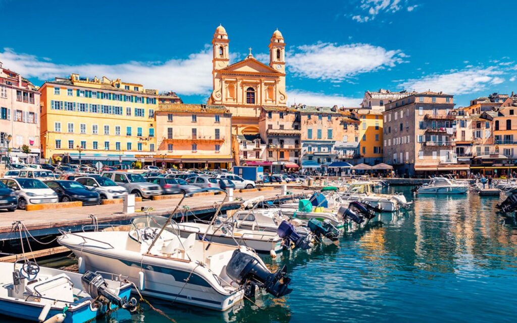 scene-dynamique-du-port-de-bonifacio-en-corse Cette image met en avant la beauté des façades colorées du port de Bonifacio, où la mer et le ciel se rejoignent pour créer une ambiance vibrante. Les bateaux alignés ajoutent au charme estival du lieu.