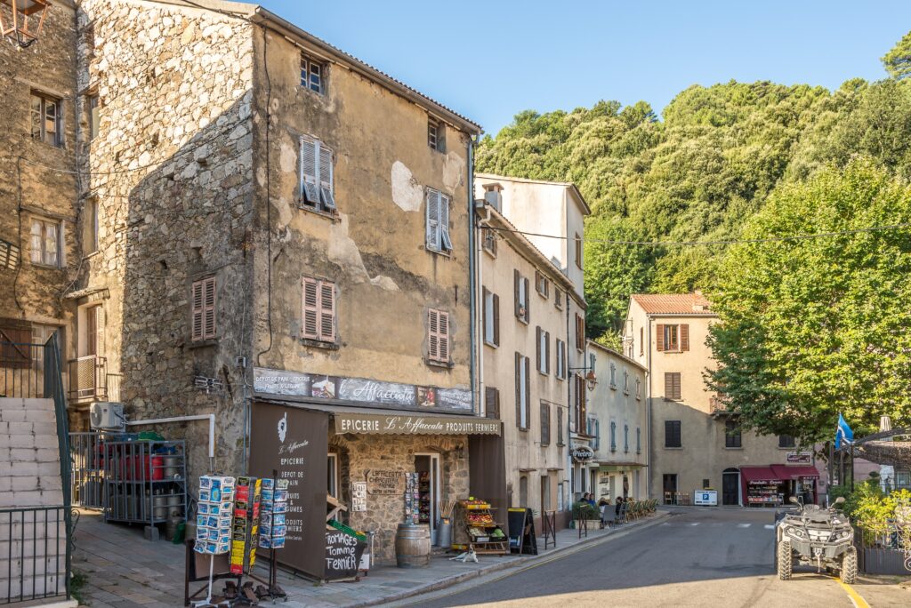 Rue pittoresque et lumineuse d'un village corse, bordée de bâtiments historiques et d'une épicerie locale, capturant l'atmosphère authentique et accueillante du Sud.