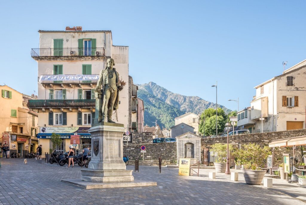 statue-historique-dans-une-place-en-corse Statue historique sur une place corse, entourée de bâtiments colorés et baignée par la lumière du soleil, avec des montagnes en arrière-plan, représentant le riche patrimoine culturel de l'île.
