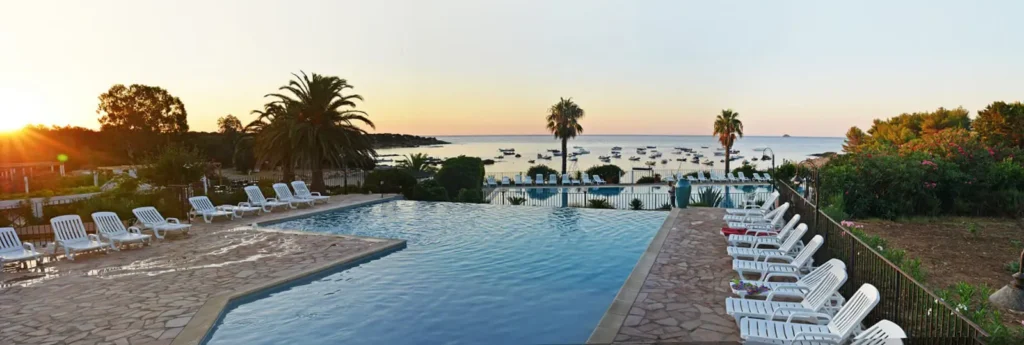 Piscine extérieure avec transats et palmiers, vue mer au coucher du soleil en Corse, ambiance camping en bord de mer.