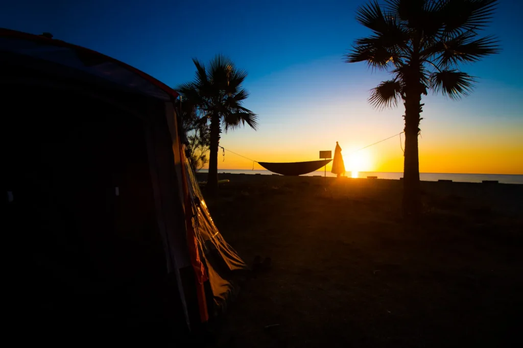 Camping+Merendella+-+Date+11072022--50 Silhouette d'une tente de camping et d'un hamac suspendu entre deux palmiers face à un coucher de soleil sur la plage au Camping Merendella.