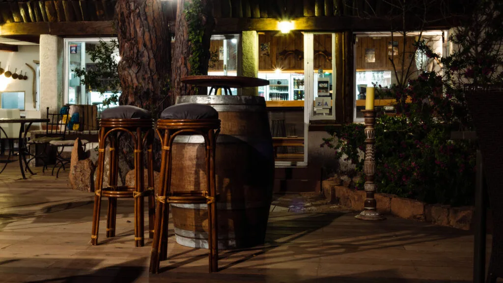Espace lounge nocturne du bar du Camping Villata avec mange-debout en tonneau et tabourets en rotin sous un éclairage tamisé.