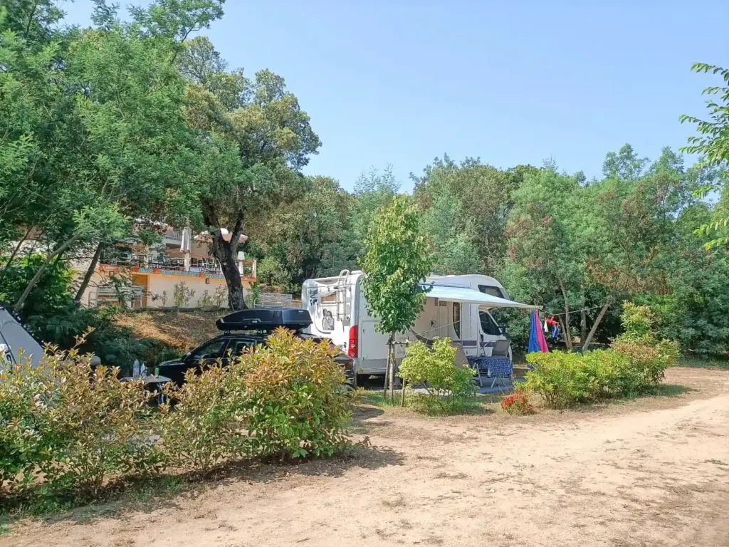 emplacement-ombragé-camping-car-tente-campervan-camping-milella Grand camping-car stationné sur un emplacement plat et herbeux avec auvent déployé, entouré d'arbres et de haies au Camping Milella.