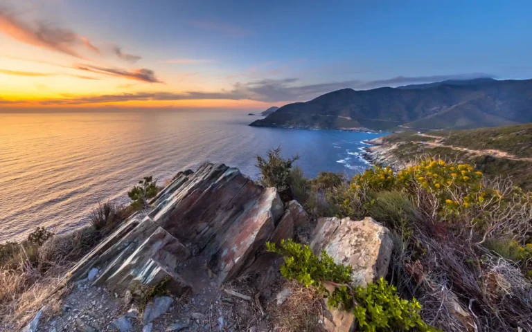 Panorama côtier au Cap Corse au coucher du soleil, pris depuis les hauteurs rocheuses. Le soleil orange se couche sur l'horizon de la mer Méditerranée, éclairant les montagnes et le maquis, avec une route côtière en contrebas.
