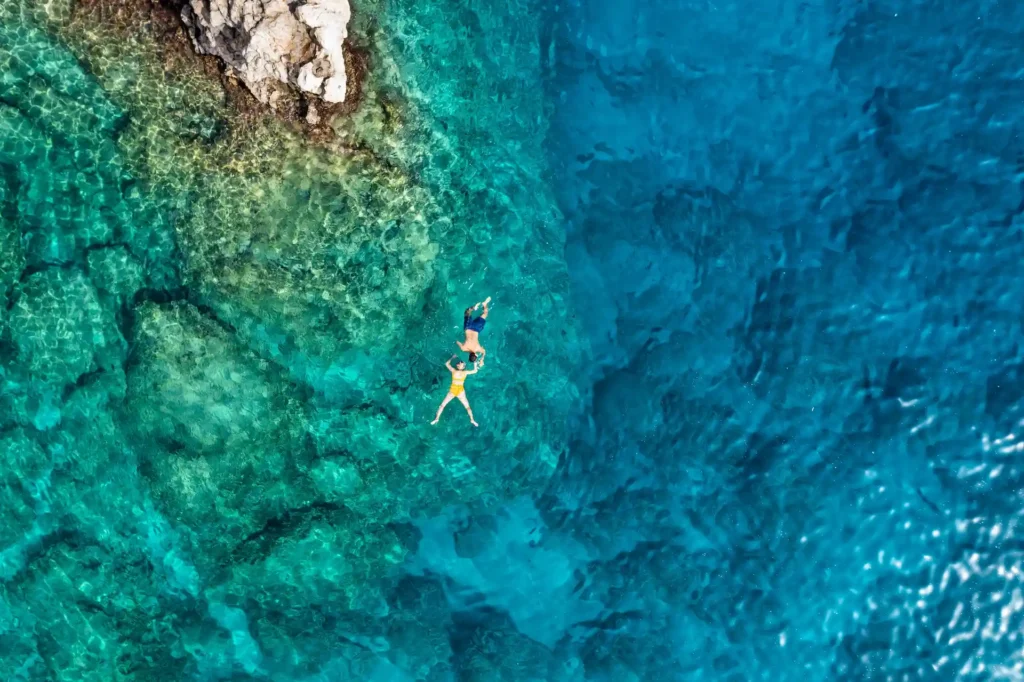 Itinéraires en bateau depuis la côte excursions aux Lavezzi, Scandola et Cap Corse Vue de drone en plongée verticale (flat lay) d'un couple se baignant main dans la main dans des eaux turquoise transparentes et limpides en Corse. Le fond marin rocheux est visible sous l'eau claire, à côté d'un rocher côtier.