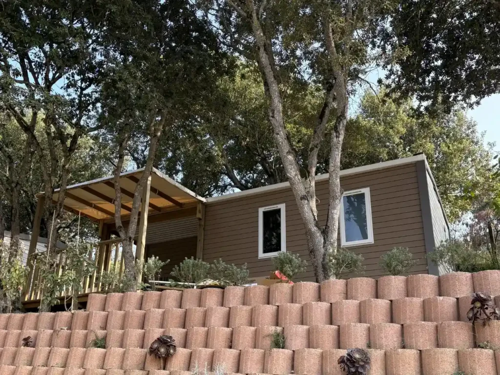 lodgia-mobilhome-confortable-camping-milella-propriano Mobil-home moderne avec bardage bois et loggia couverte, installé sur un terrain en terrasse soutenu par un mur de soutènement végétalisé.