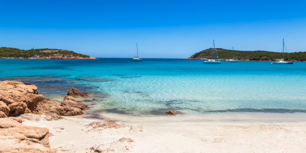 Vue panoramique d'une baie sauvage en Corse avec catamarans au mouillage et plage de sable blanc accessible à pied depuis le camping.