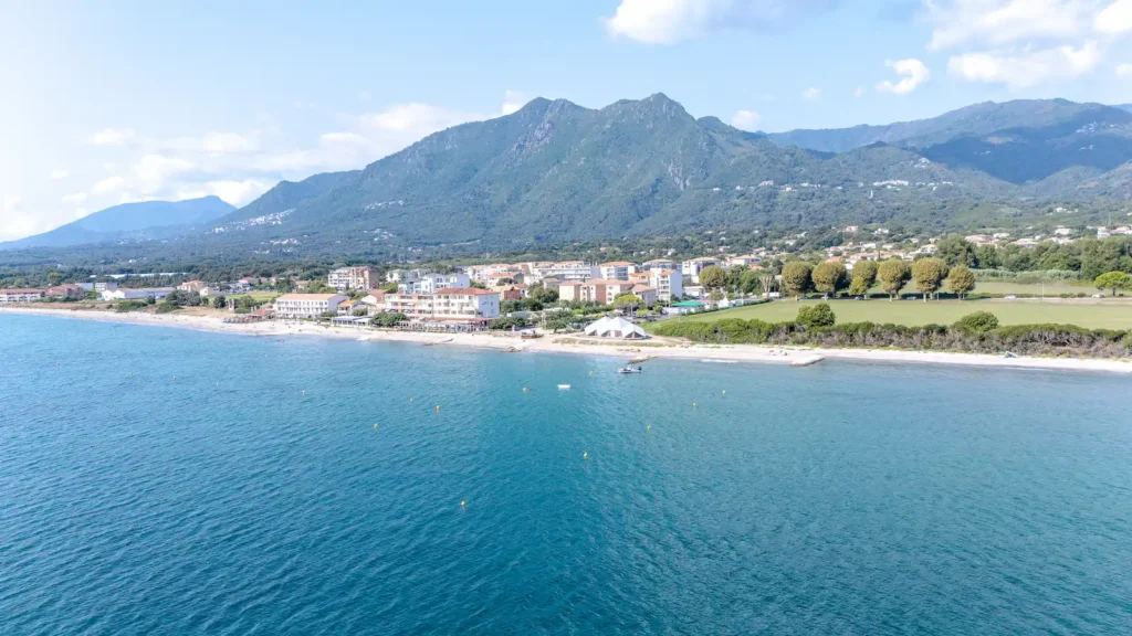 vue-moriani-plage-costa-verde-aventure Vue aérienne d'une station balnéaire corse avec ses plages de sable fin, ses infrastructures de camping et ses montagnes verdoyantes en arrière-plan.
