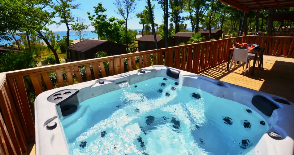 hebergement-insolite-corse-bord-de-mer Jacuzzi privatif bouillonnant sur la terrasse en bois d'un hébergement insolite en Corse avec vue sur la mer et la plage à travers les arbres.